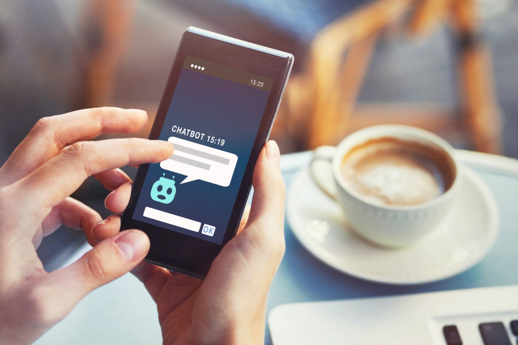 Chatbot auf einem Smartphone, welches von einer Person gehalten wird, die an einem Tisch mit Kaffetasse und Laptop sitzt.